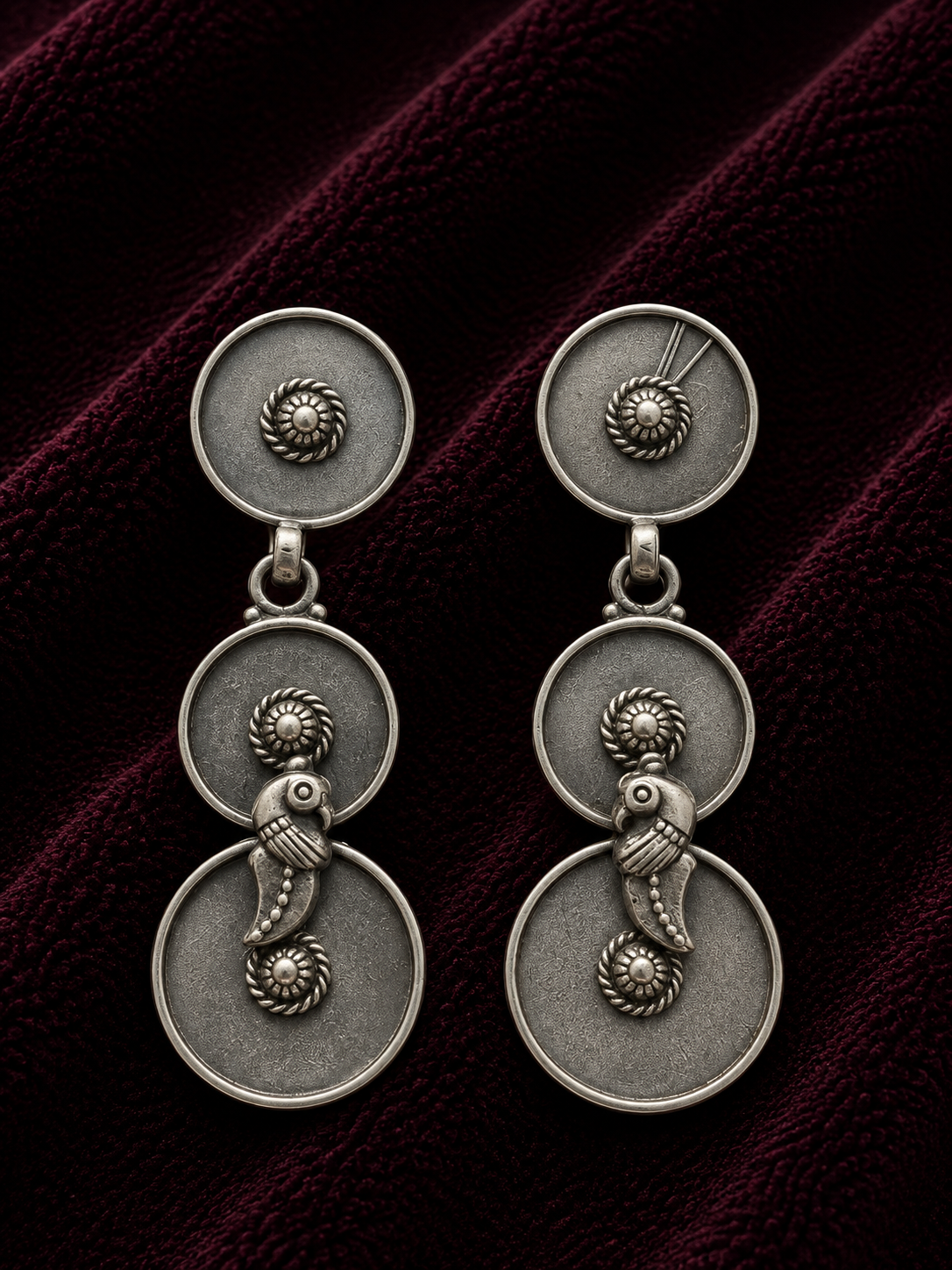 A pair of antique silver dangle earrings with circular stacked discs and a small fish motif, displayed on a dark burgundy velvet fabric background.