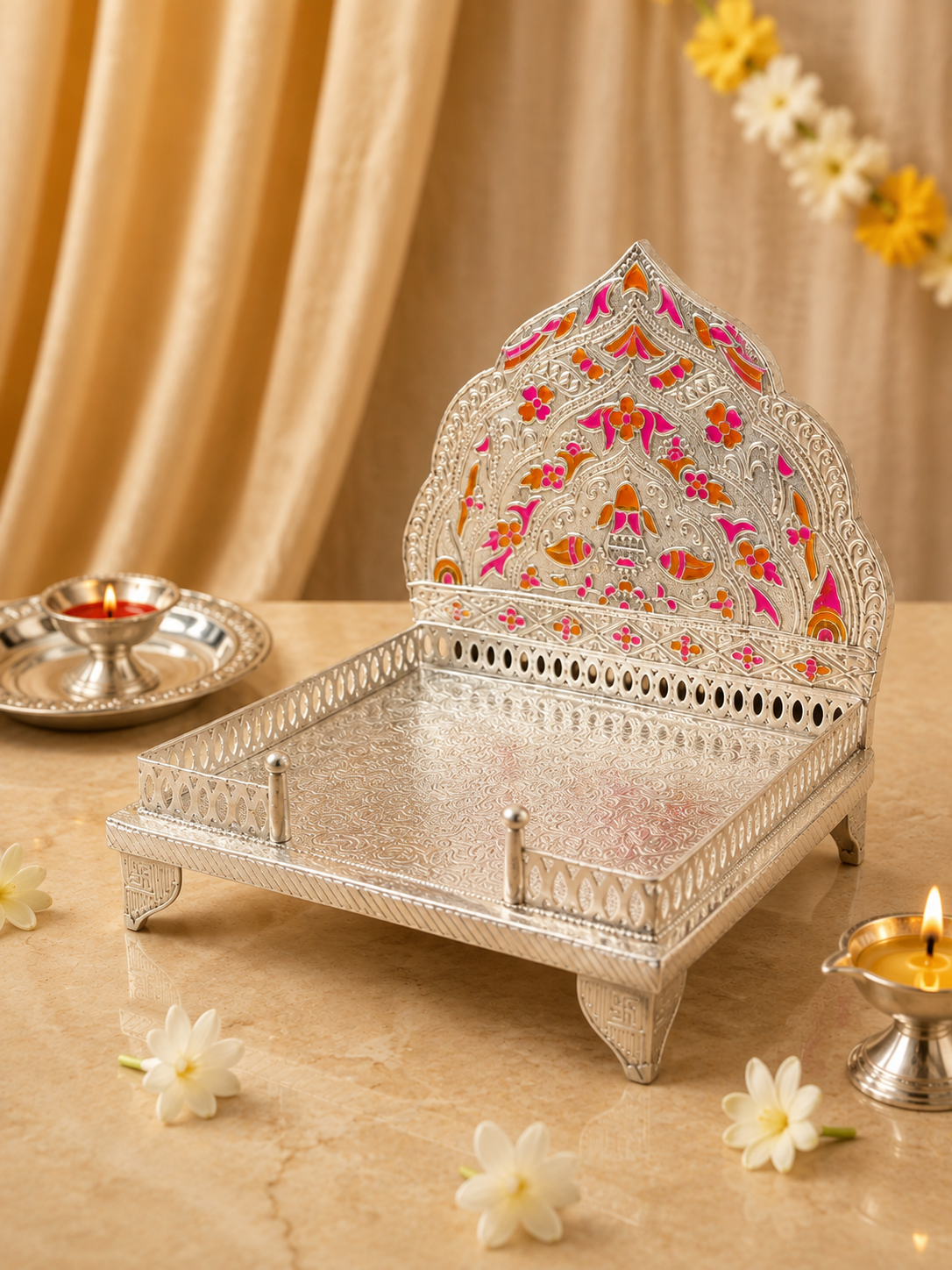 An ornate silver-toned decorative tray stand with a tall arched backrest, decorated with colorful floral motifs in pink, orange, and yellow. The stand is placed on a beige marble-like surface with lit diya candles, white flowers, and draped beige fabric in the background.