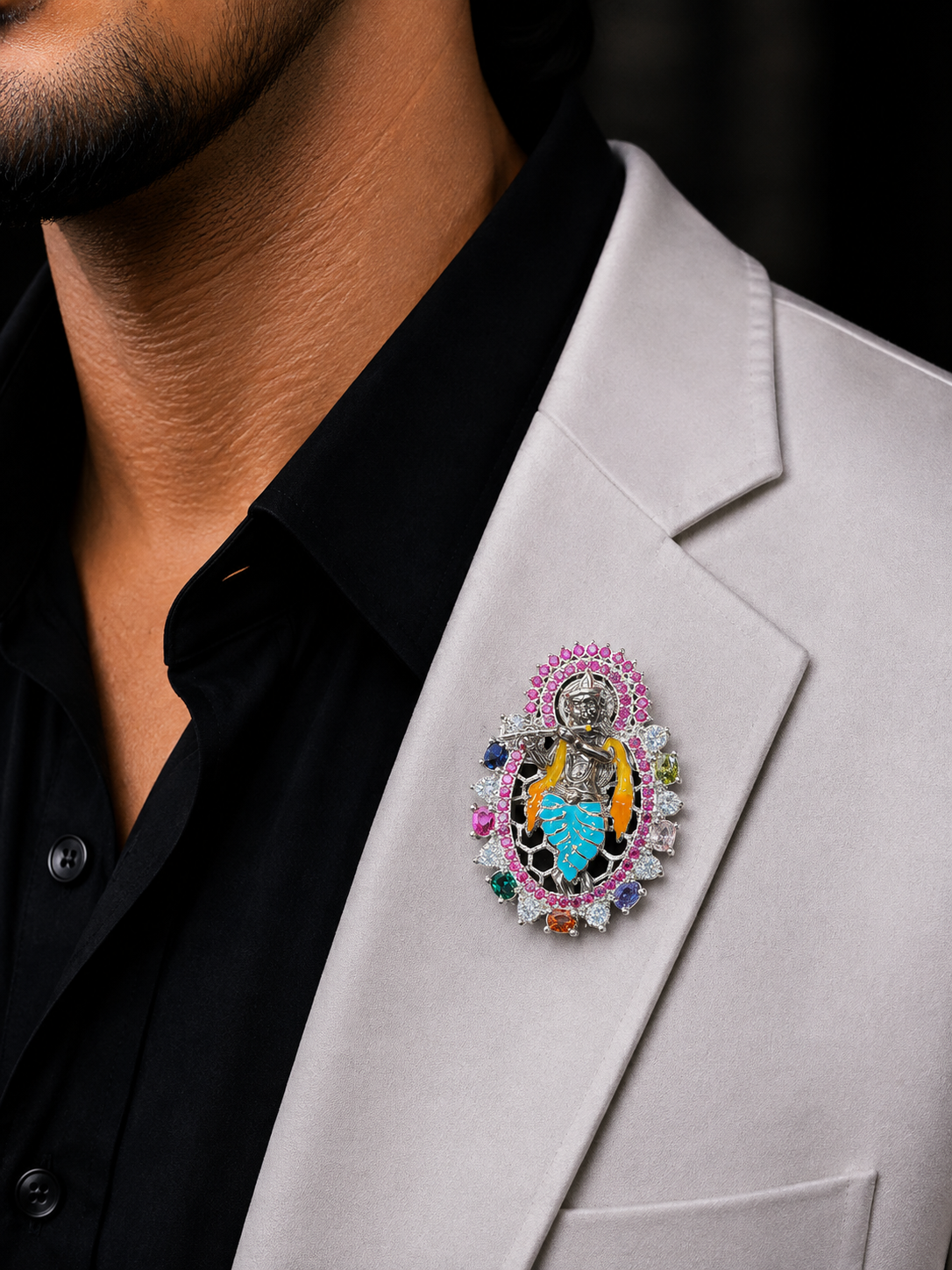 A close-up of a man wearing a black collared shirt and a light gray blazer, featuring a highly detailed, colorful religious brooch pinned to his lapel. The brooch depicts a silver-toned figure surrounded by vibrant pink, blue, yellow, and green gemstone-like accents.