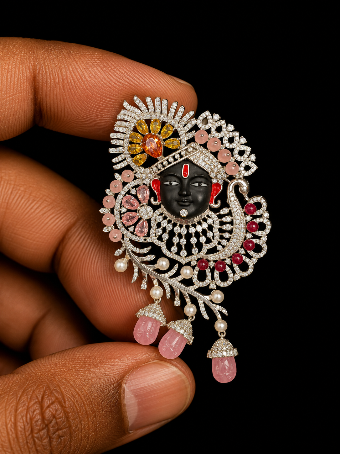 A close-up of a hand holding an ornate silver-toned jewelry piece featuring a black face, pink gemstones, red stones, and white pearl-like accents against a black background.
