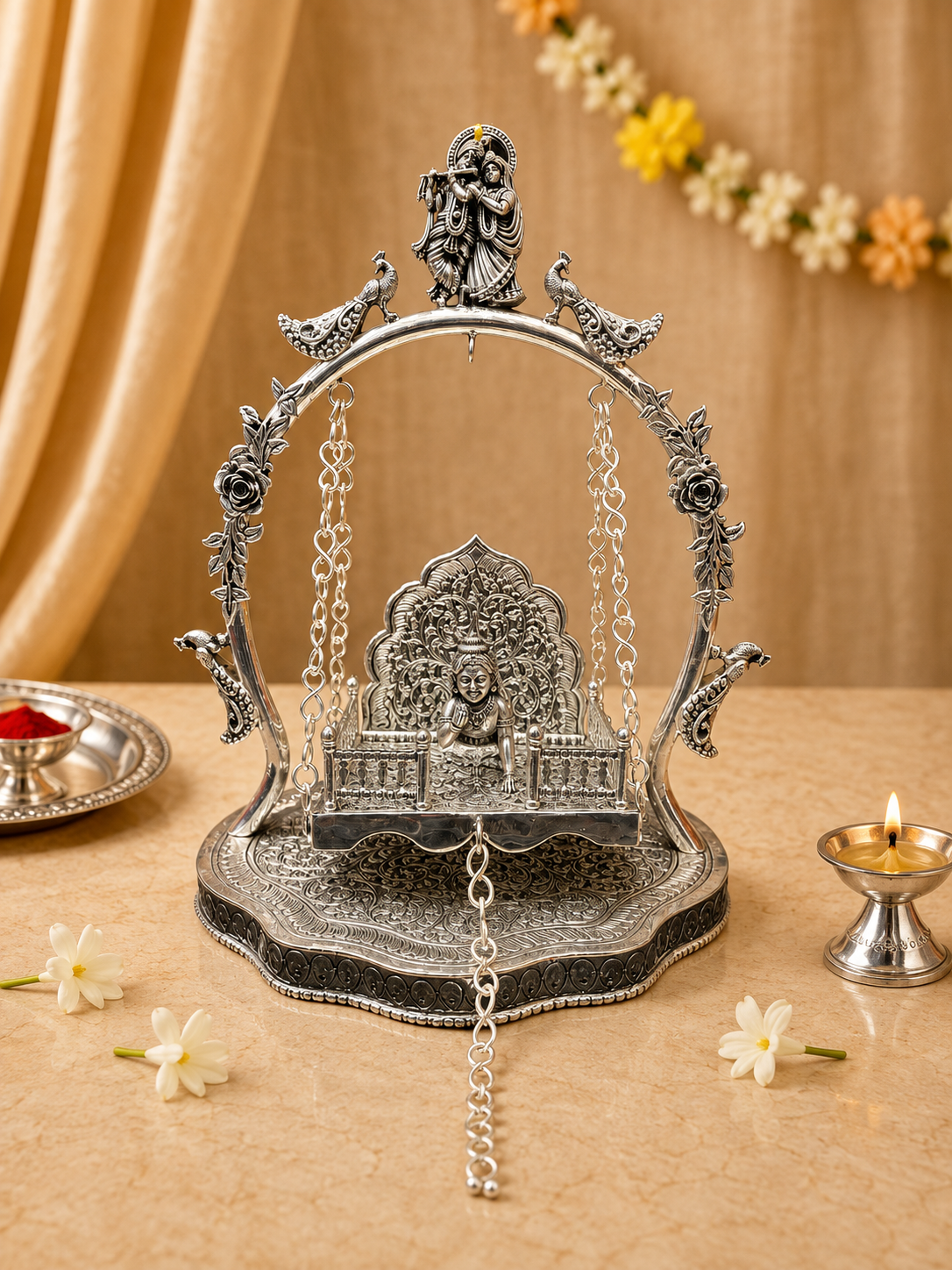 An ornate silver-toned swing-style decorative idol stand with a seated deity figure, floral and bird motifs, and hanging chains, displayed on a beige surface with warm draped fabric, flowers, and small lit lamps in the background.