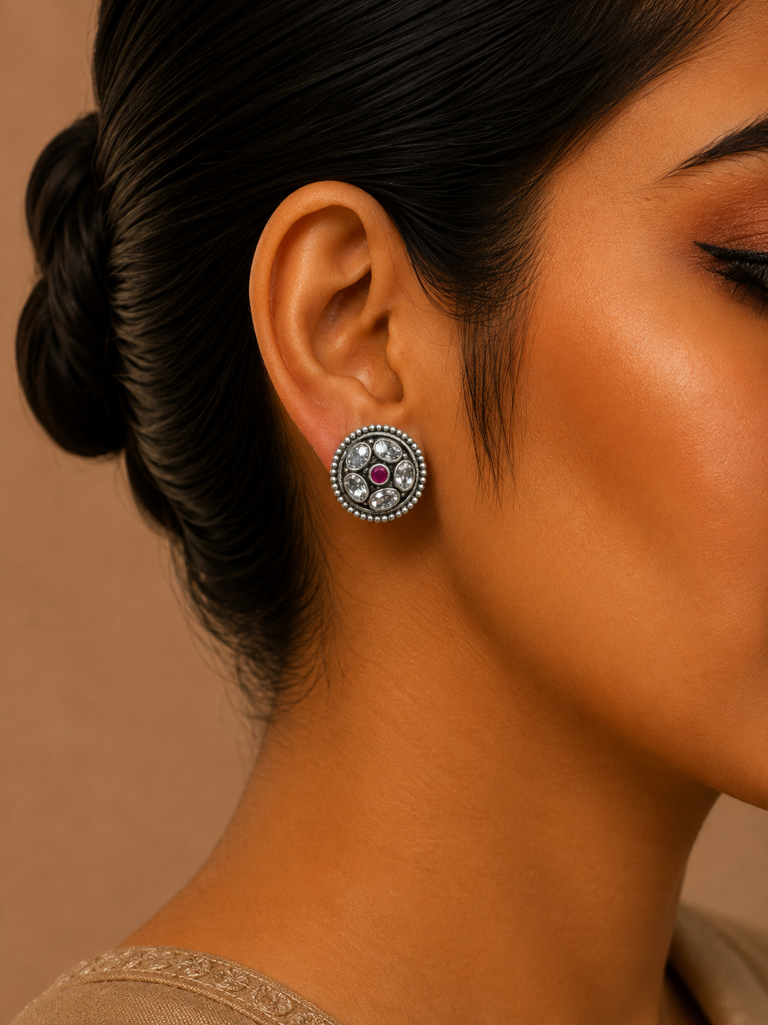 A close-up side profile of a woman wearing a round silver-toned stud earring with a central dark red gemstone and surrounding clear stones, against a warm beige background.