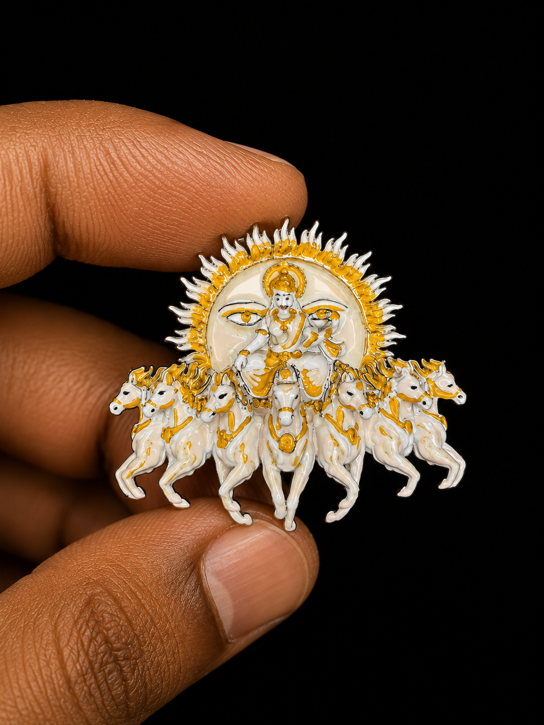 A hand holding a detailed white and gold religious pin depicting Lord Ganesha on a horse with the other three horses around him, set against a solid black background.