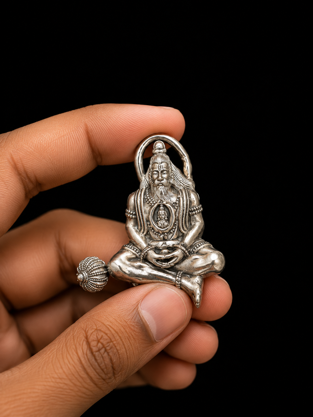 A hand holding a small silver-toned metal figurine of a seated deity with intricate carved details, set against a solid black background.