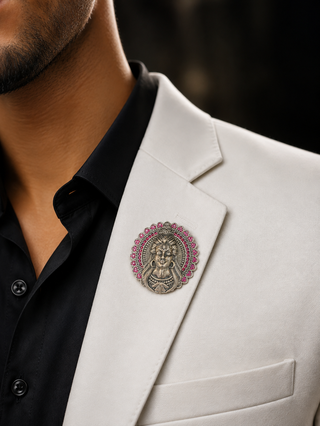A close-up of a man wearing a white suit jacket over a black collared shirt, featuring a circular silver-toned brooch with pink gemstone accents on the lapel.