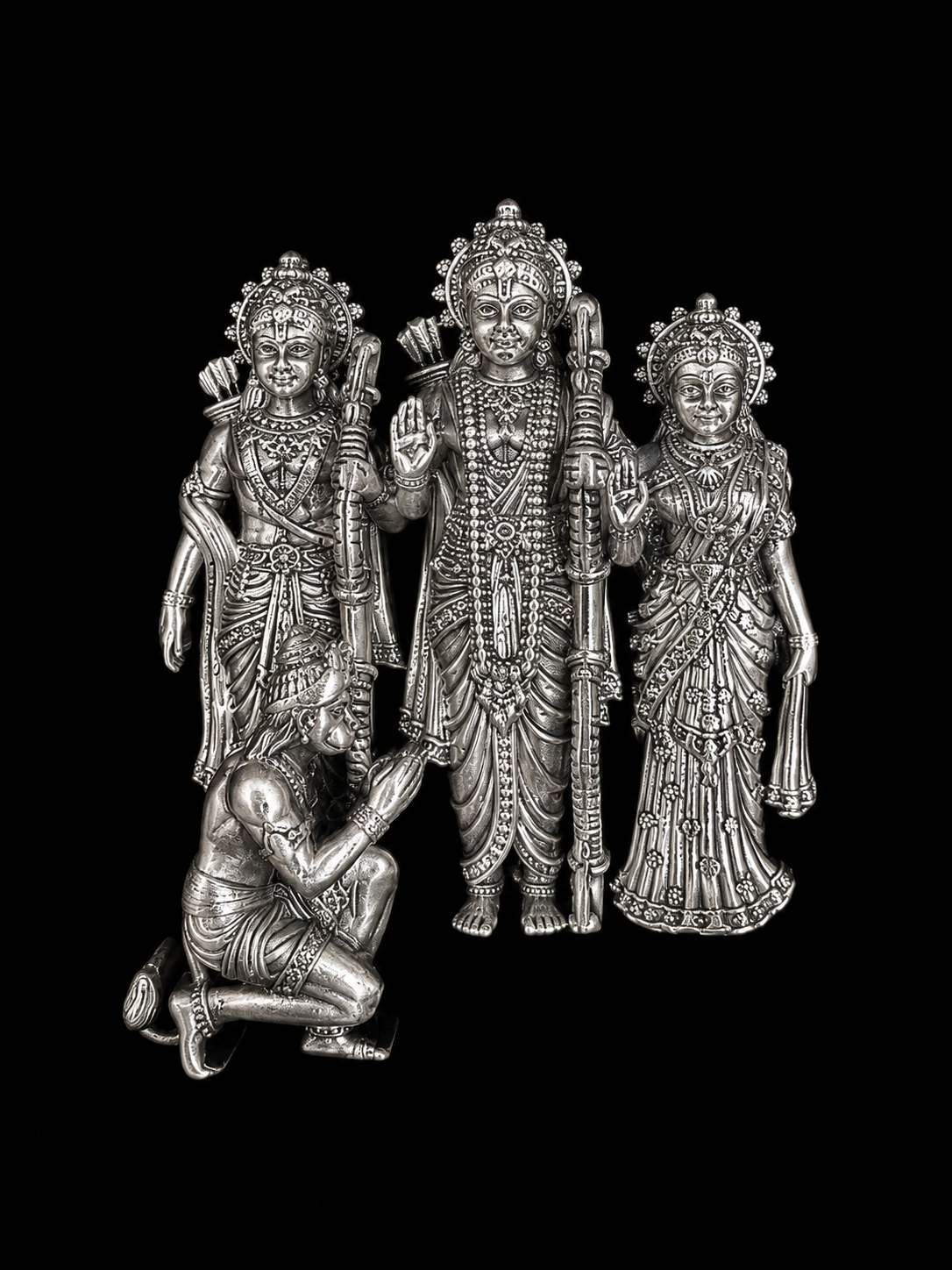 An ornate silver-toned metal figurine depicting a Hindu family scene with three standing figures and one kneeling figure, shown against a solid black background.