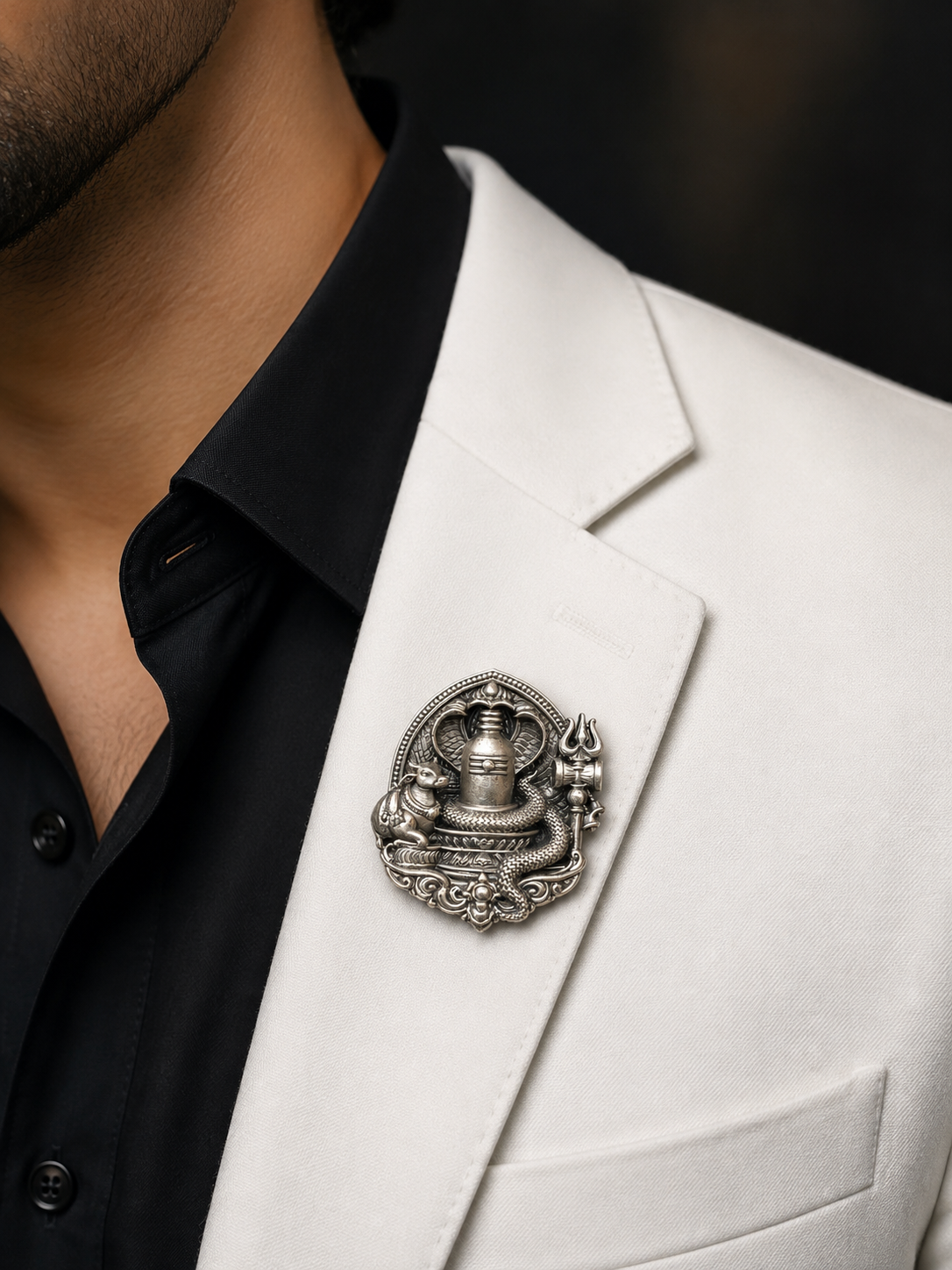A close-up of a man wearing a white blazer over a black collared shirt, featuring a detailed silver-toned metal brooch pinned to the lapel.