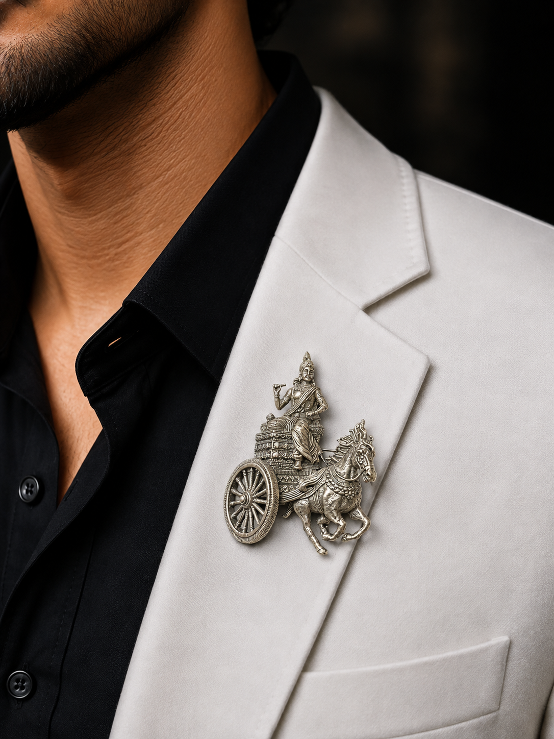 A close-up of a man wearing a detailed silver-toned brooch shaped like a chariot with a deity figure on horseback, pinned to his white blazer over a black shirt.