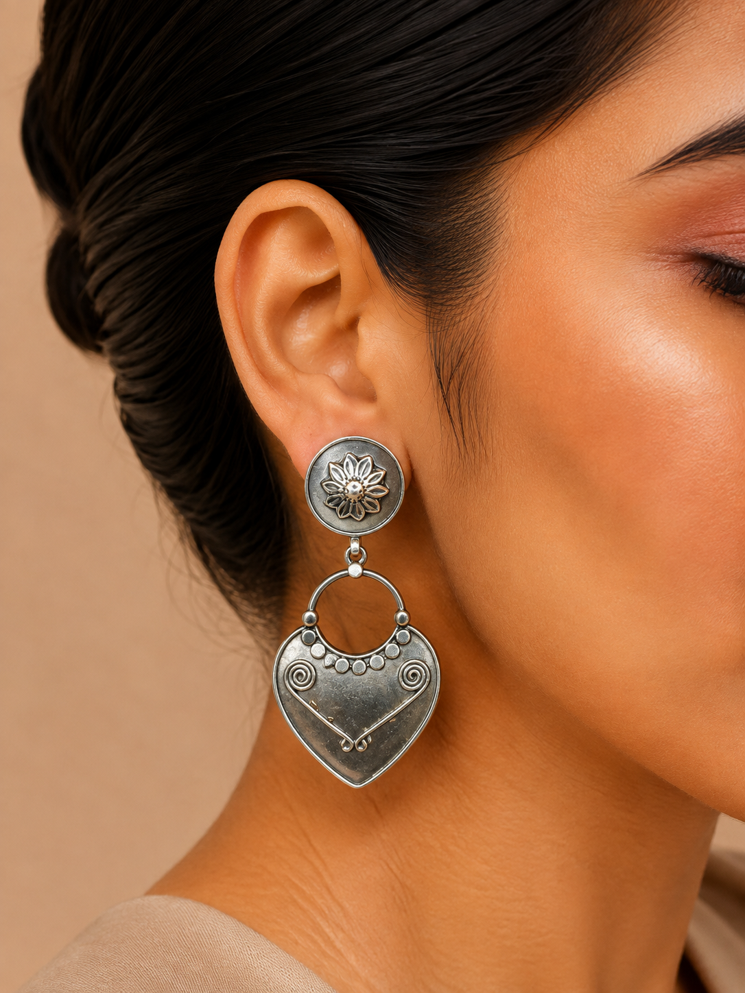 A close-up of a model's ear wearing a silver-toned double drop earring. The earring features a circular stud with a lotus flower motif and a dangling heart-shaped charm with engraved swirls and a small key detail, set against a warm beige background.